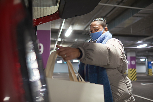 Woman in protective mask packing shopping bags into her car while standing in underground parking