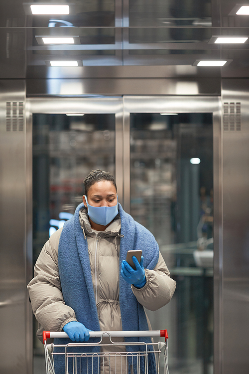 Young woman in protective mask standing in elevator with shopping cart and making selfie on the phone