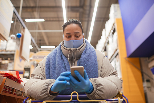 Young woman in protective mask and gloves typing a message on mobile phone while standing with shopping cart in supermarket