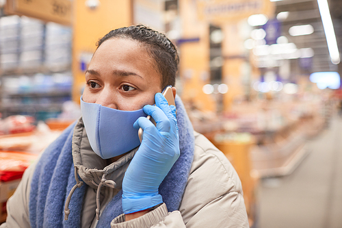 Young woman in protective mask talking on mobile phone while standing in supermarket