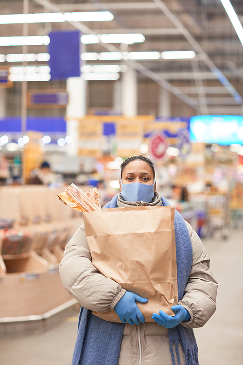 Young woman in mask holding shopping bags and looking at camera while standing in supermarket during pandemic