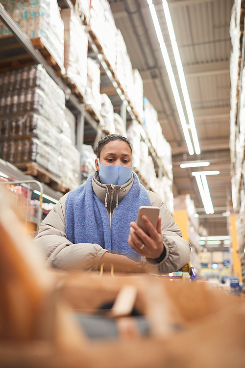 Young woman in protective mask making photo on her mobile phone while doing shopping in supermarket