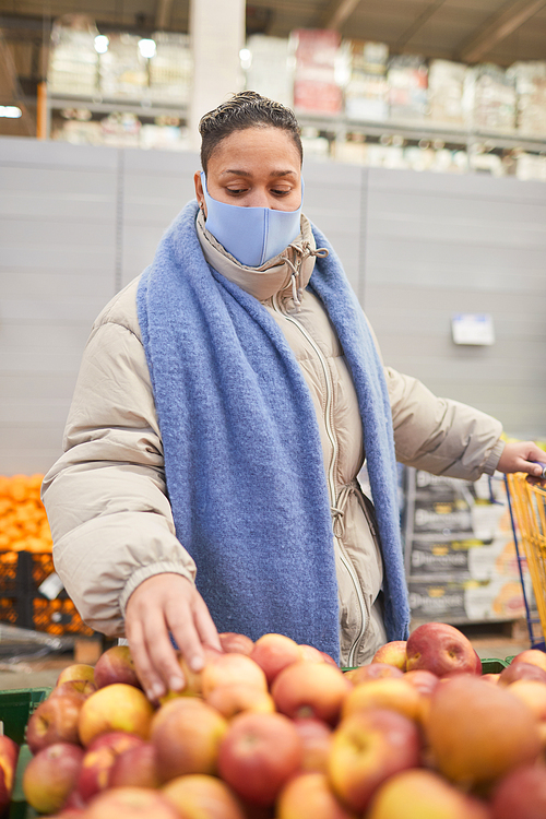 Young woman in protective mask doing shopping in supermarket during pandemic choosing fresh apples