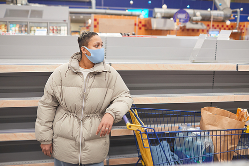 Young woman in protective mask standing with shopping cart in supermarket and doing shopping