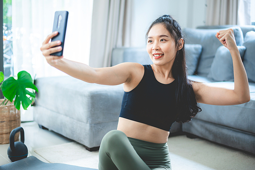 Portrait of cheerful young Asian woman in sportswear showing biceps and muscles sitting on yoga mat and taking a selfie from smartphone at home after practicing yoga and exercise
