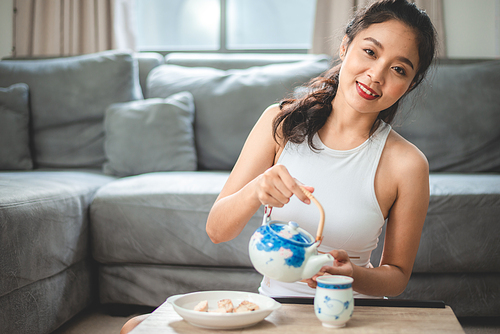 Portrait of smiling young Asian beautiful woman sitting on floor near table enjoying healthy breakfast while pouring tea in cup from teapot and looking at camera at home