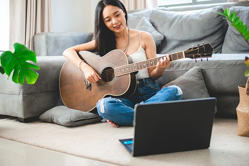 Young beautiful Asian woman guitarist holding and ready to play instrument guitar while learning and practicing notes and music with the help of online training and coaching using laptop at home