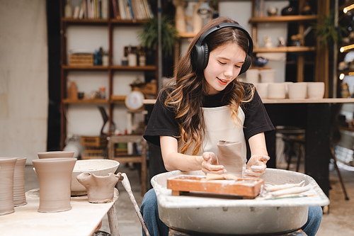 Beautiful young woman holding pottery instrument for scraping, smoothing, shaping and sculpting. Lady siting on bench with pottery wheel and making clay pot