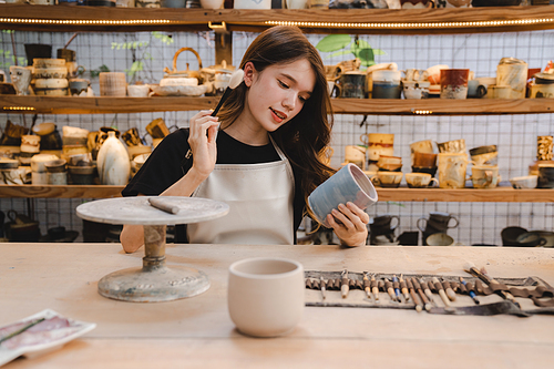 Beautiful young woman holding pottery instrument for scraping, smoothing, shaping and sculpting. Lady siting on bench with pottery wheel and making clay pot