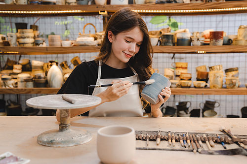 Beautiful young woman holding pottery instrument for scraping, smoothing, shaping and sculpting. Lady siting on bench with pottery wheel and making clay pot