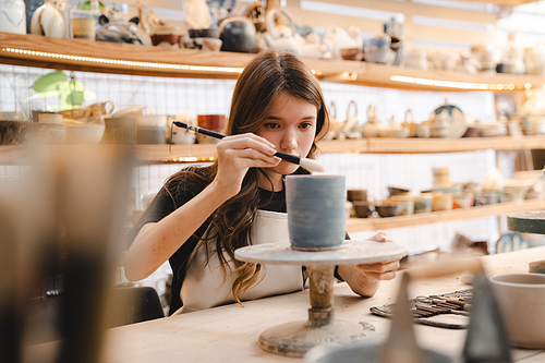 Beautiful young woman holding pottery instrument for scraping, smoothing, shaping and sculpting. Lady siting on bench with pottery wheel and making clay pot