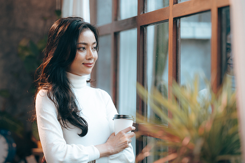 Smiling and successful young female Asian entrepreneur working on laptop while taking a break by sipping coffee and tea and looking away while thinking about new strategy and relaxing