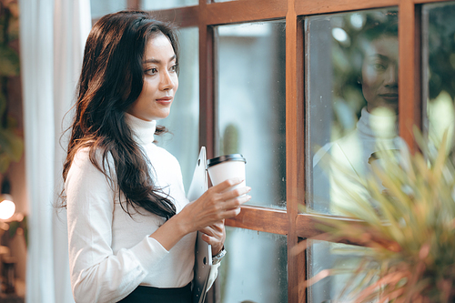 Smiling and successful young female Asian entrepreneur working on laptop while taking a break by sipping coffee and tea and looking away while thinking about new strategy and relaxing