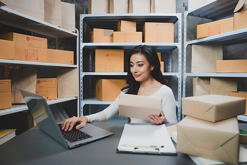 Smiling and successful young female Asian entrepreneur working and holding digital tablet with pen and working in warehouse and storage room while shipping and taking parcels and creating stock