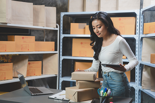 Smiling and successful young female Asian entrepreneur working and holding digital tablet with pen and working in warehouse and storage room while shipping and taking parcels and creating stock