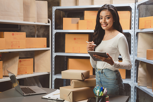 Smiling and successful young female Asian entrepreneur working and holding digital tablet with pen and working in warehouse and storage room while shipping and taking parcels and creating stock