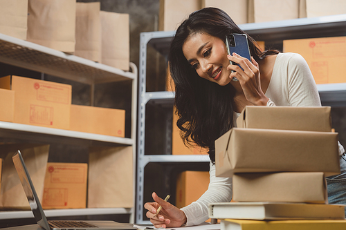 Smiling and successful young female Asian warehouse manager and worker working in storage house while checking package and parcel and placing in shelf and rack