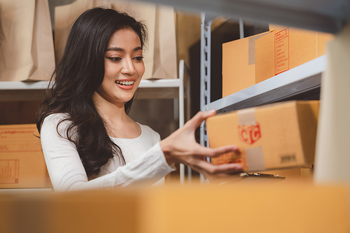 Smiling and successful young female Asian warehouse manager and worker working in storage house while checking package and parcel and placing in shelf and rack