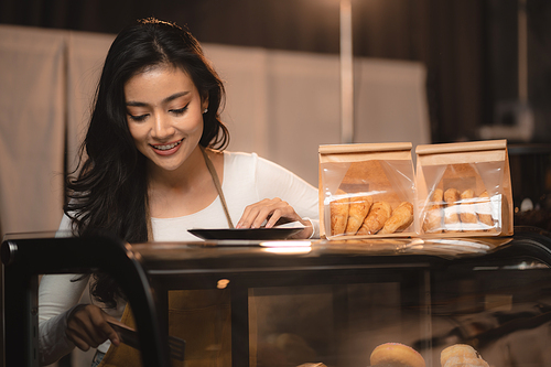 Bakery and coffee cafe business shop concept, Asian female wearing aprons, happily smiling to serving breakfast baked bread, beautiful woman staff service customer to delivery a food