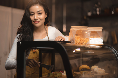 Bakery and coffee cafe business shop concept, Asian female wearing aprons, happily smiling to serving breakfast baked bread, beautiful woman staff service customer to delivery a food