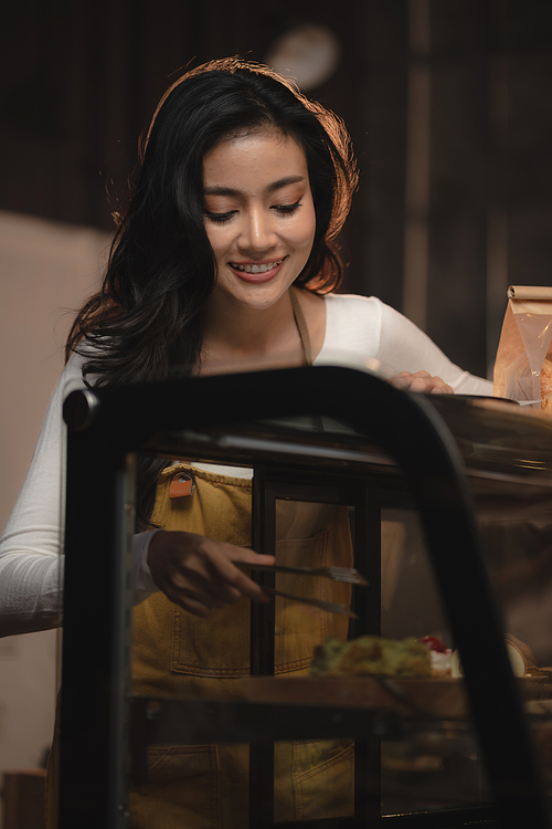 Bakery and coffee cafe business shop concept, Asian female wearing aprons, happily smiling to serving breakfast baked bread, beautiful woman staff service customer to delivery a food