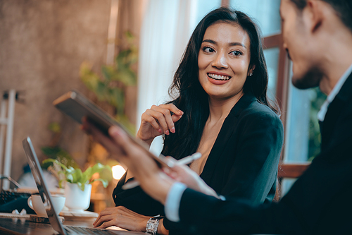 Smiling and laughing young Asian successful business woman discussing work with male colleague and partner in modern office while using digital tablet