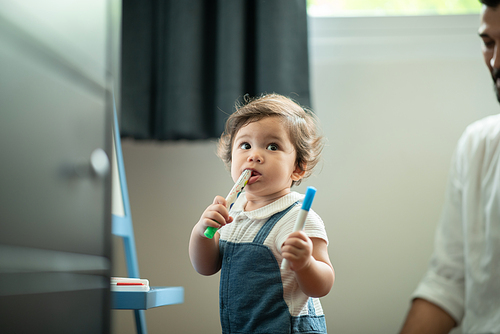 Cute little toddler baby boy playing and chewing marker pen with father while looking away and thinking at home in living room while learning