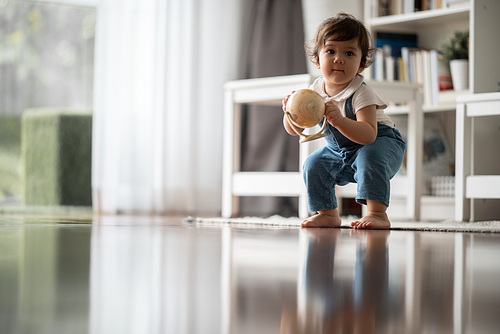 Cute little baby boy toddler sitting on wooden floor playing with globe ball while looking away and thinking of broken toy in living room at home