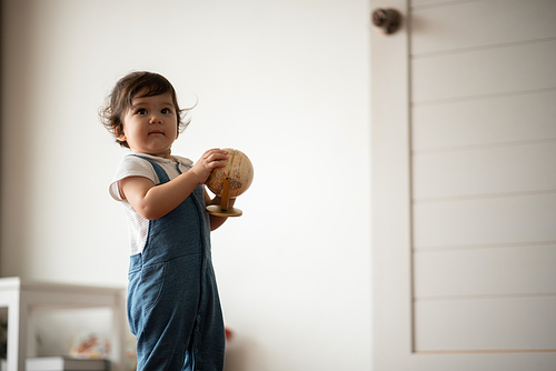 Cute little baby boy toddler sitting on wooden floor playing with globe ball while looking away and thinking of broken toy in living room at home