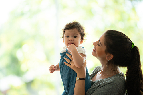 Portrait of playful and naughty cute little baby toddler playing and enjoying while in the arms of young mother hugging at home looking at camera