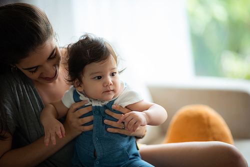 Portrait of playful and naughty cute little baby toddler playing and enjoying while in the arms of young mother hugging at home looking at camera