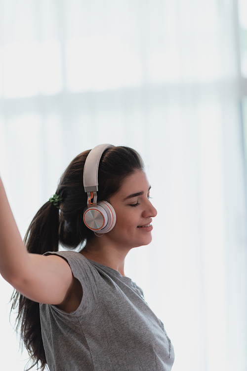 Portrait of young and carefree young woman in casual clothing listening to music using wireless bluetooth headphones dancing and enjoying at home with eyes closed and relaxing