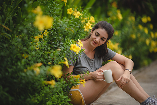 Cheerful and beautiful young carefree woman relaxing and breathing fresh air while sitting in balcony with nature view and drinking morning coffee and beverage and looking at scenic view