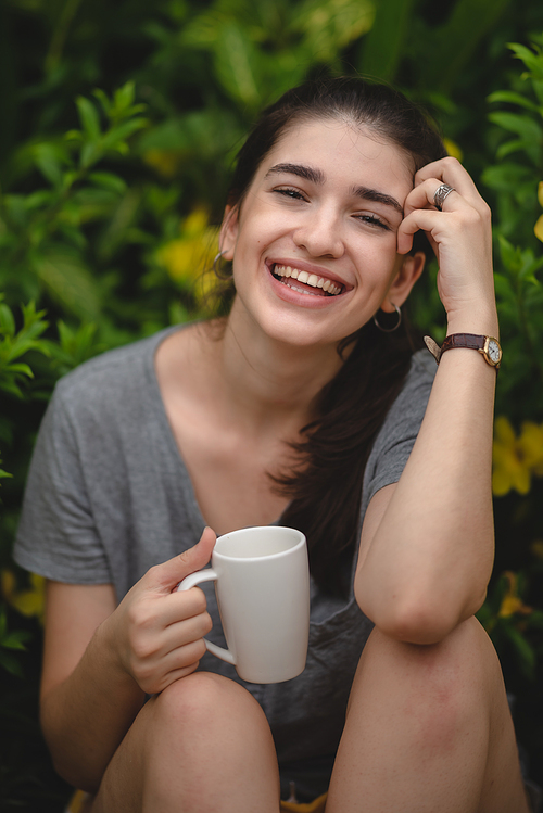 Cheerful and beautiful young carefree woman relaxing and breathing fresh air while sitting in balcony with nature view and drinking morning coffee and beverage and looking at scenic view