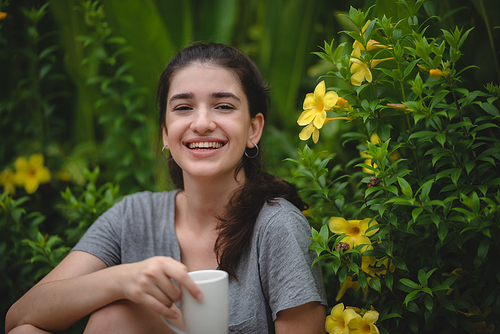 Cheerful and beautiful young carefree woman relaxing and breathing fresh air while sitting in balcony with nature view and drinking morning coffee and beverage and looking at scenic view