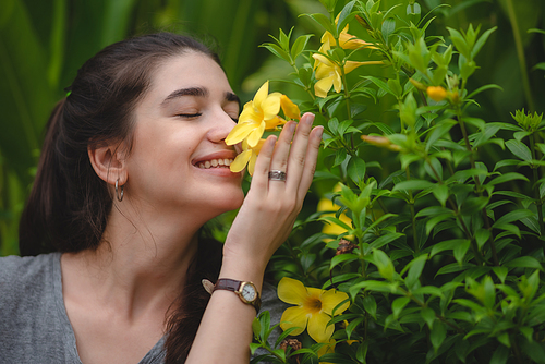 Beautiful young woman with toothy smile feeling and smelling beautiful yellow flower while in nature enjoying and relaxing with closed eyes