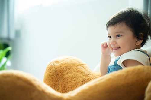 Cute little playful girl child learning to walk and stand by taking support of couch at home while looking away mischievously