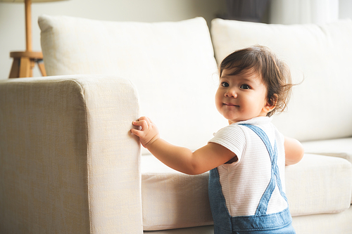 Cute little playful girl child learning to walk and stand by taking support of couch at home while looking away mischievously