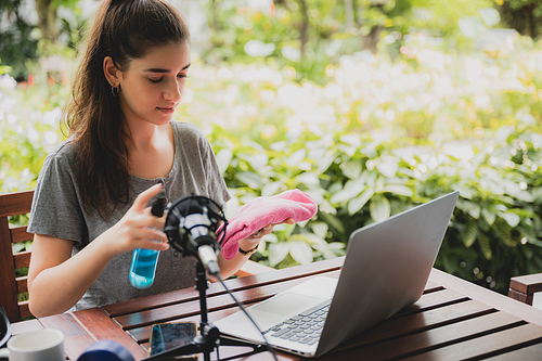 Young female vlogger with mic and dumbbells holding a cloth and spray bottle to clean laptop screen or recording a video to give information about a product while sitting at home