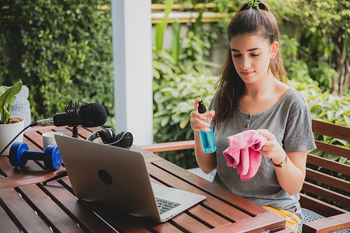 Young female vlogger with mic and dumbbells holding a cloth and spray bottle to clean laptop screen or recording a video to give information about a product while sitting at home