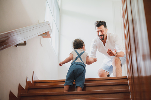 Handsome young father playing with little child daughter who is climbing stairs at home and man motivating her to come upstairs