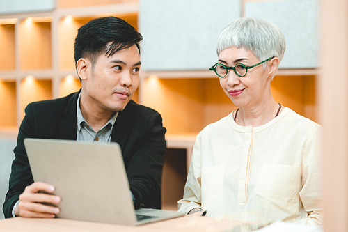 Young Asian business man in formal clothing showing something on digital tablet to senior female boss and manager wearing eyeglasses while in a meeting and training on technology in modern office