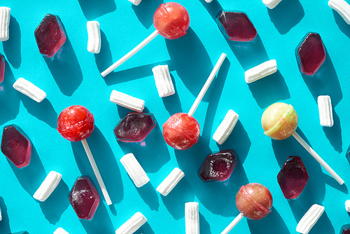 Colorful composition of sweets for every taste: appetizing lollipops, light and airy marshmallows and candied fruit jelly lying in disorder against blue background, directly above view