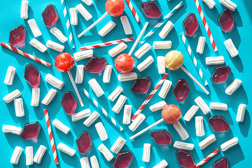 Composition of striped drinking straws, bright lollipops, airy marshmallows and pieces of candied fruit jelly lying in a certain order against blue background, directly above view