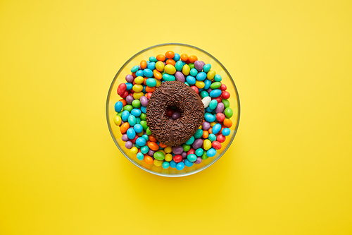 High angle view of jelly beans and chocolate doughnut with sprinkles in glassy bowl on yellow background