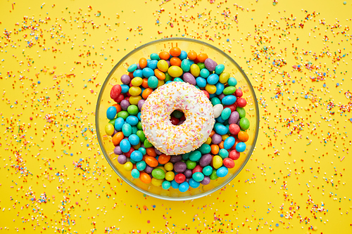 Directly above view of glassy bowl full of candies and doughnut, yellow background with sweet sprinkles