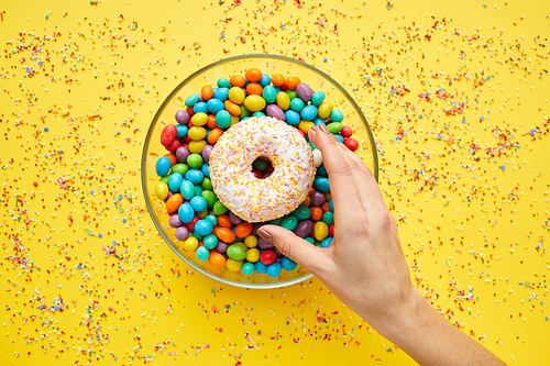 Top view of unrecognizable woman reaching hand to take sweet doughnut from bowl of candies, yellow background