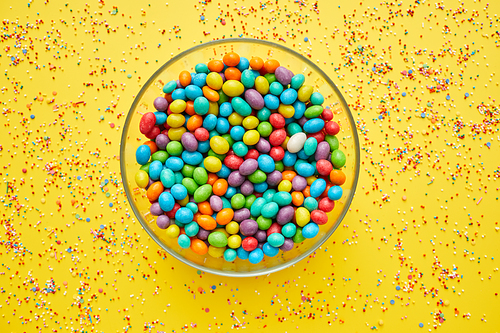 Glassy bowl full of colorful sweet beans on yellow background with splattering sprinkles