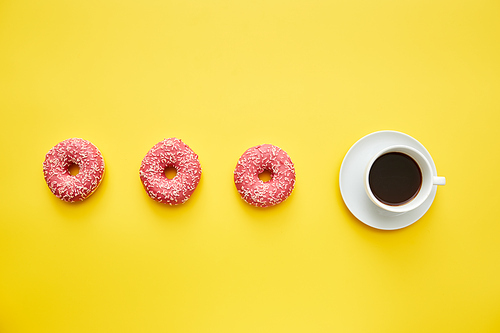 Cup of black coffee and pink doughnuts with sprinkles in line on yellow background, calories concept