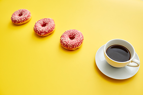 Breakfast concept: close-up of full coffee cup on plate and doughnuts with pink topping placed in a row, yellow background